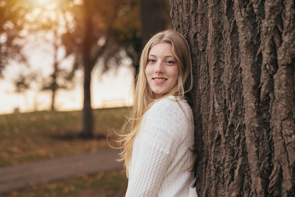 Person in white knit sweater leans against tree trunk in autumn park during golden sunset.