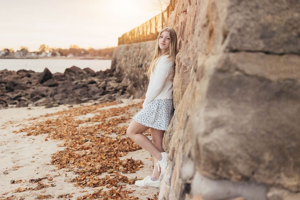 A person in a white dress leans against a stone wall on a rocky beach at sunset.