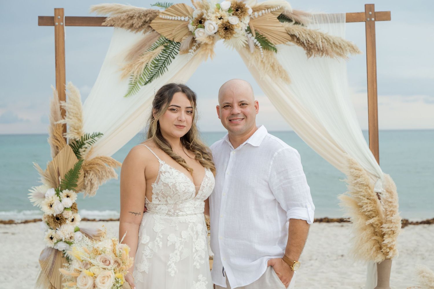 Couple standing together under a boho beach wedding arch decorated with pampas grass and white fabric by the ocean.