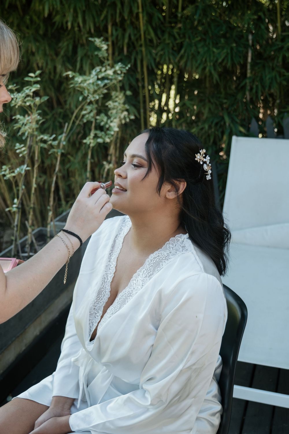 A person in a white lace top getting makeup applied while sitting outdoors near bamboo plants.