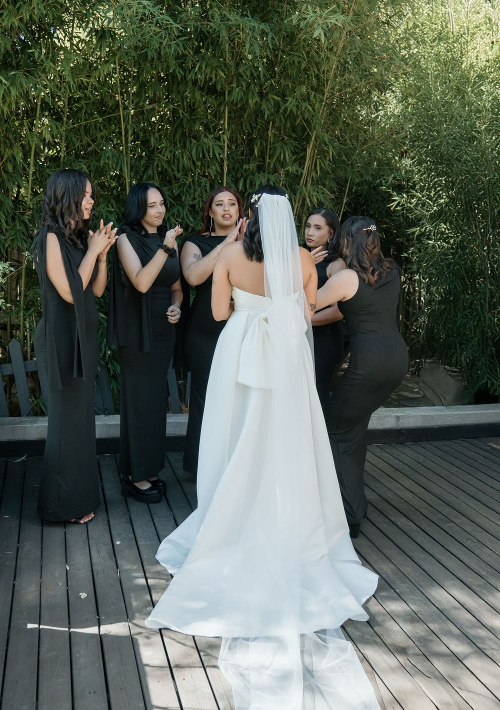 Bride in flowing white gown shares a candid moment with bridesmaids wearing black dresses on wooden deck surrounded by bamboo.