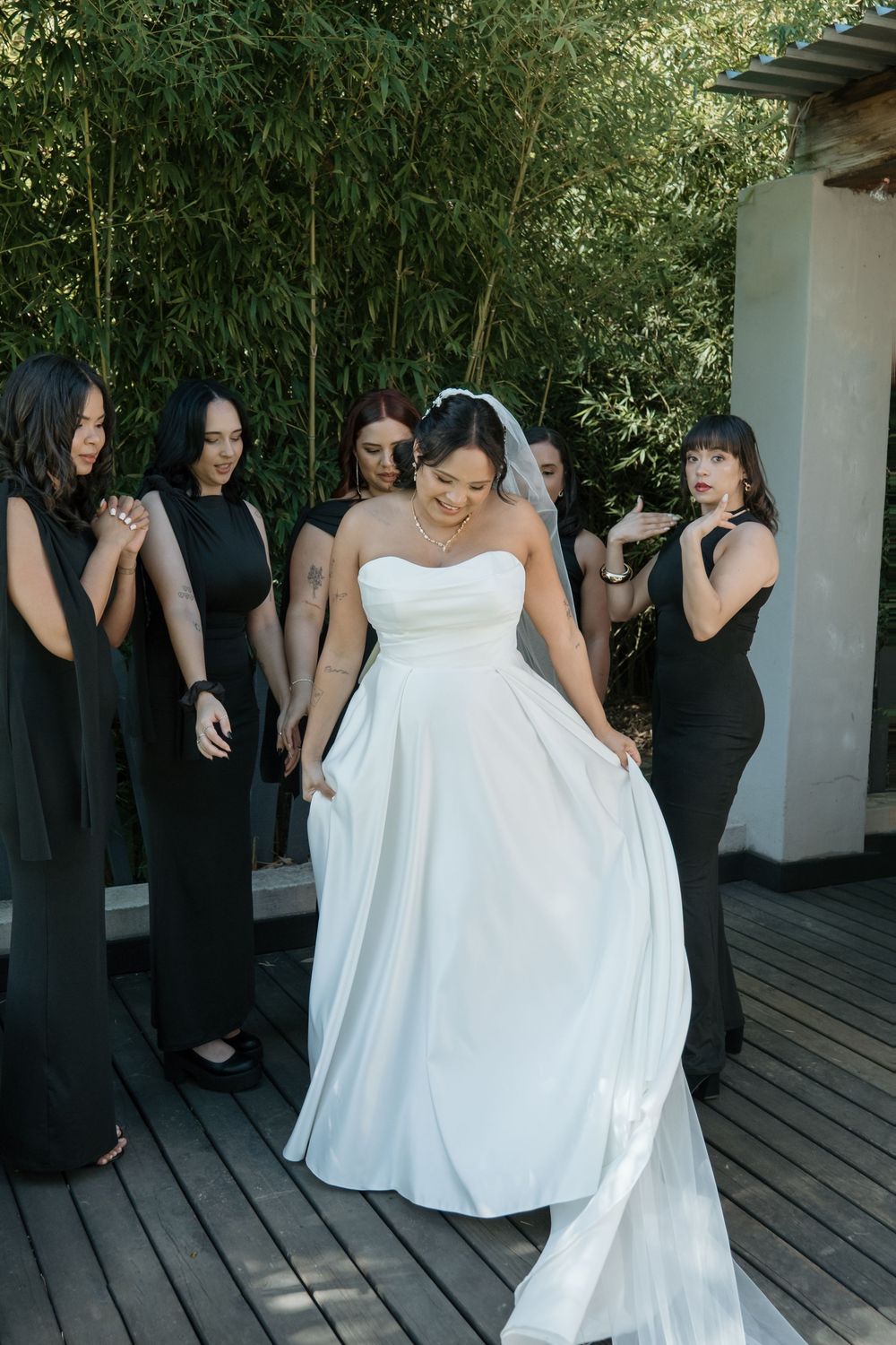 A bride in a flowing white gown twirls on a wooden deck surrounded by bridesmaids wearing elegant black dresses.