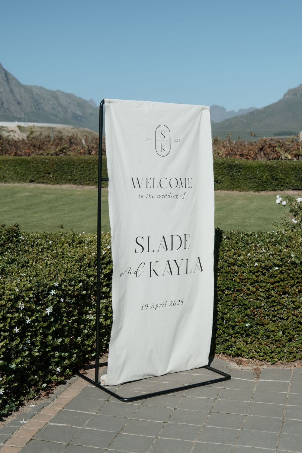 A series of white wedding welcome signs with elegant text displayed along a stone pathway against mountain and garden backdrops.