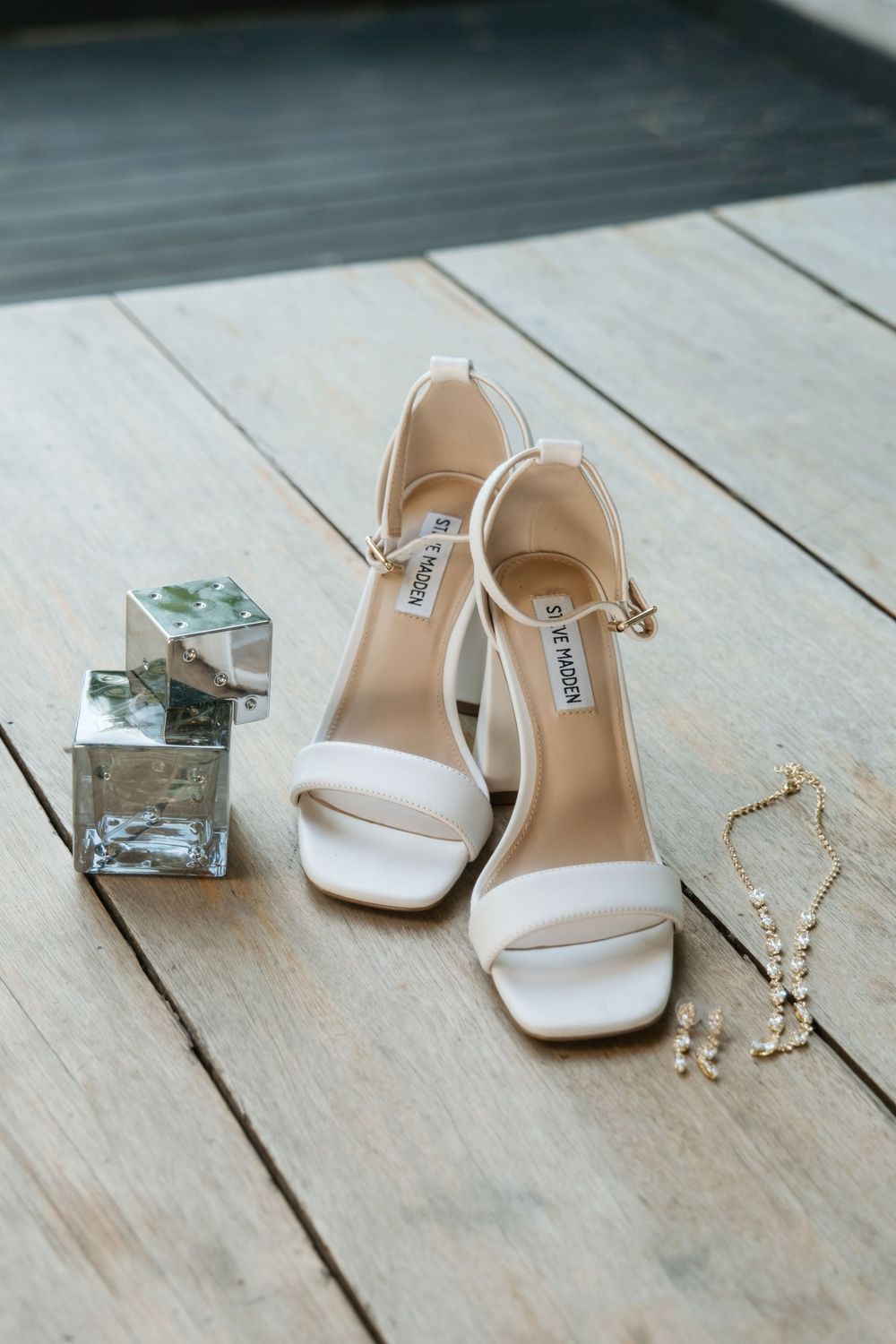Elegant white wedding heels displayed with crystal perfume bottles and pearl earrings on a rustic wooden floor.