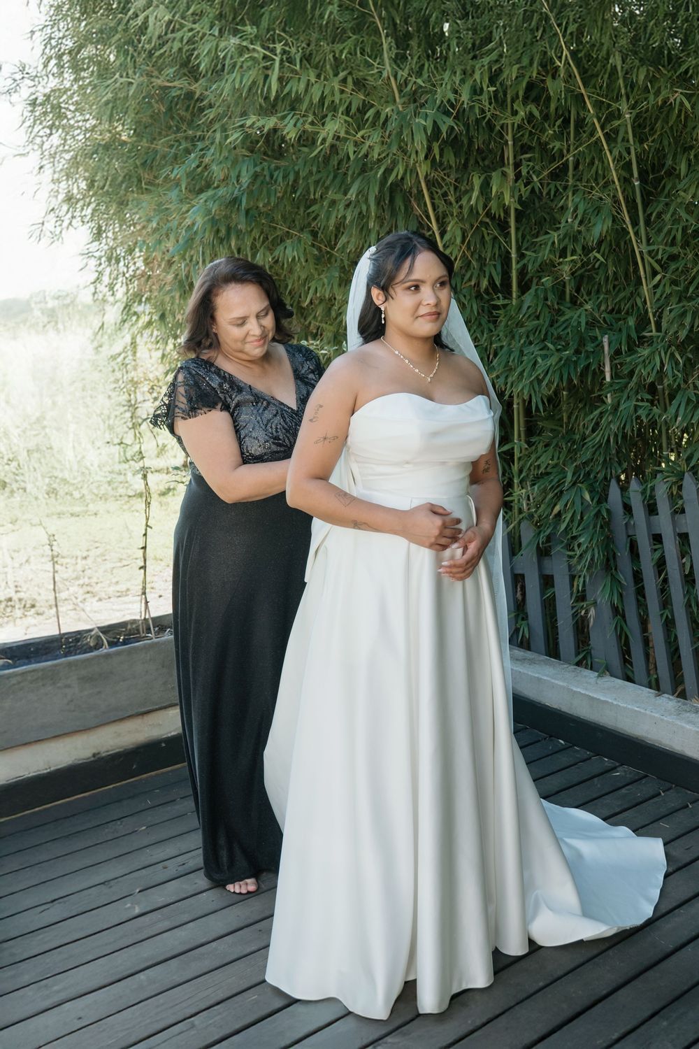 A person in a black formal dress helps adjust a white wedding gown on a deck with bamboo plants in the background.