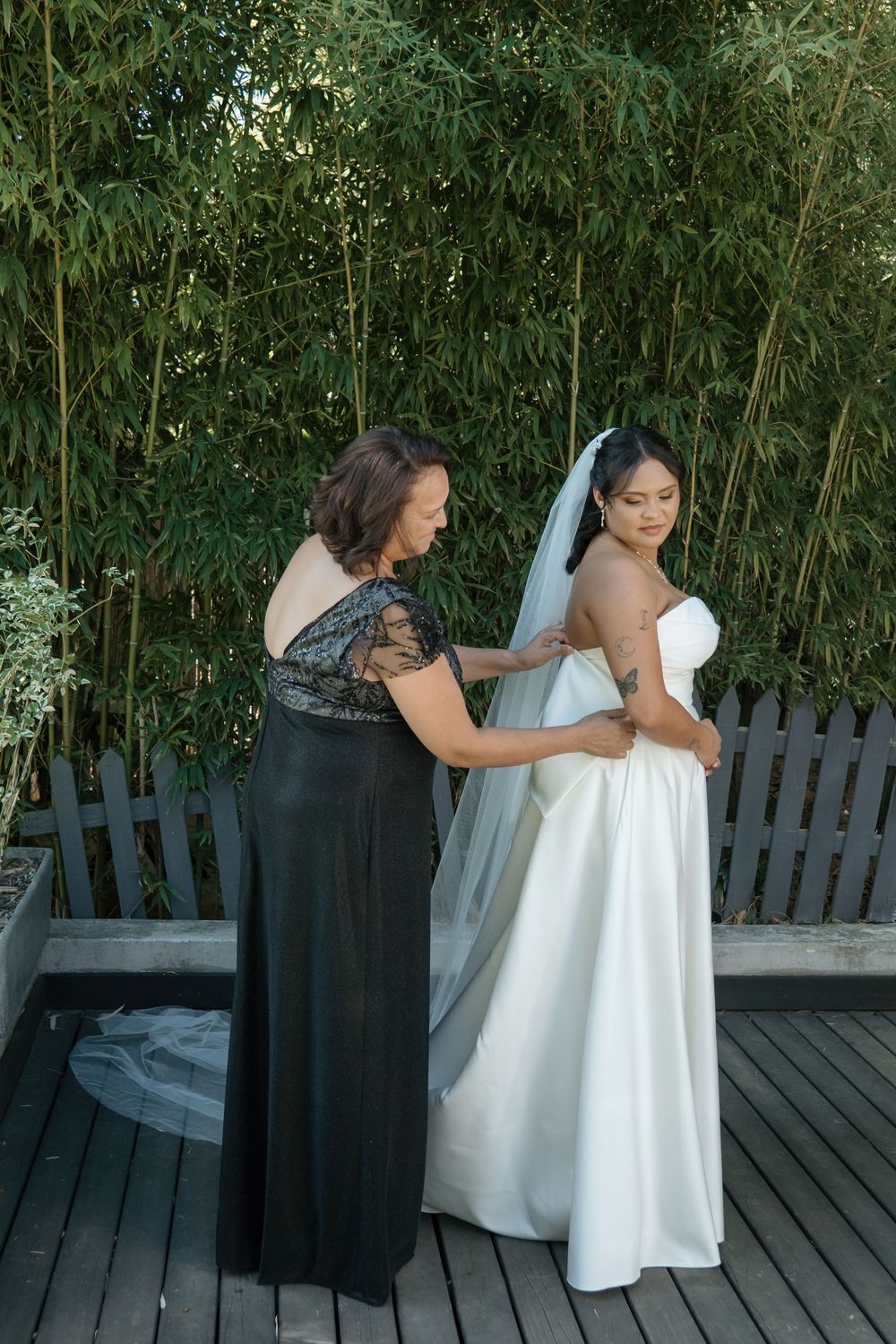 A wedding preparation moment with a black-gowned figure helping adjust a flowing white bridal gown on a wooden deck.