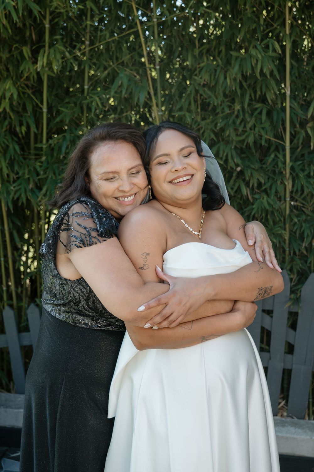 Two people share a warm embrace and joyful smiles while standing in front of green foliage on a sunny day.