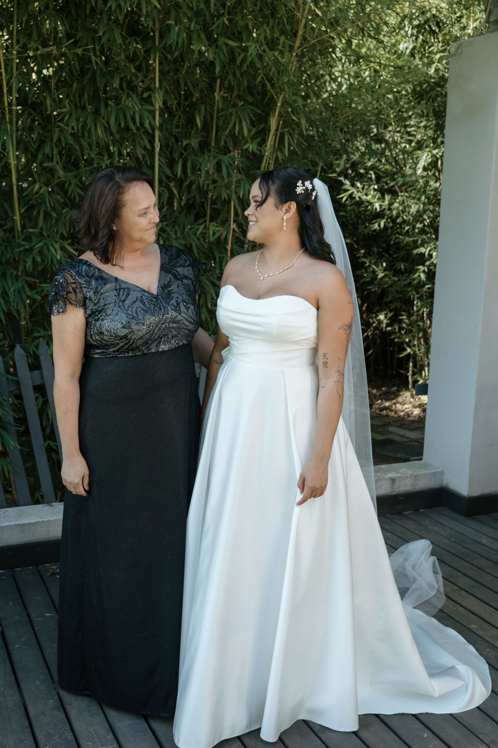 Two people celebrate a wedding moment outdoors in black and white dresses on a wooden deck surrounded by greenery.
