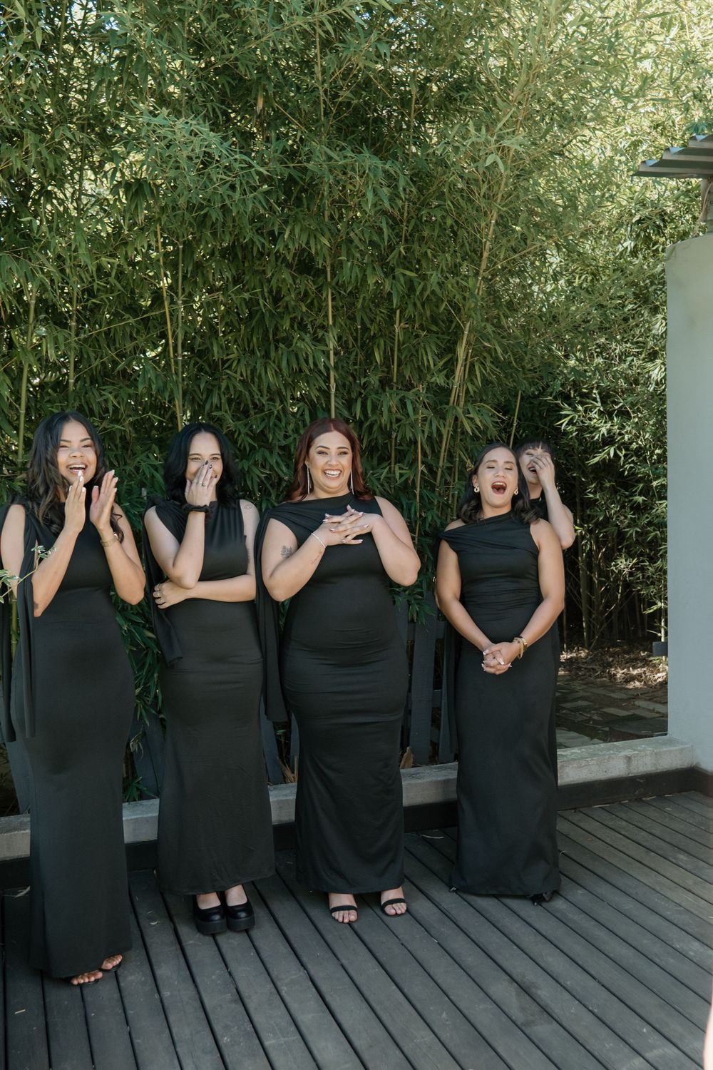 A group of people in coordinated black formal dresses pose together on a wooden deck with bamboo plants in the background.