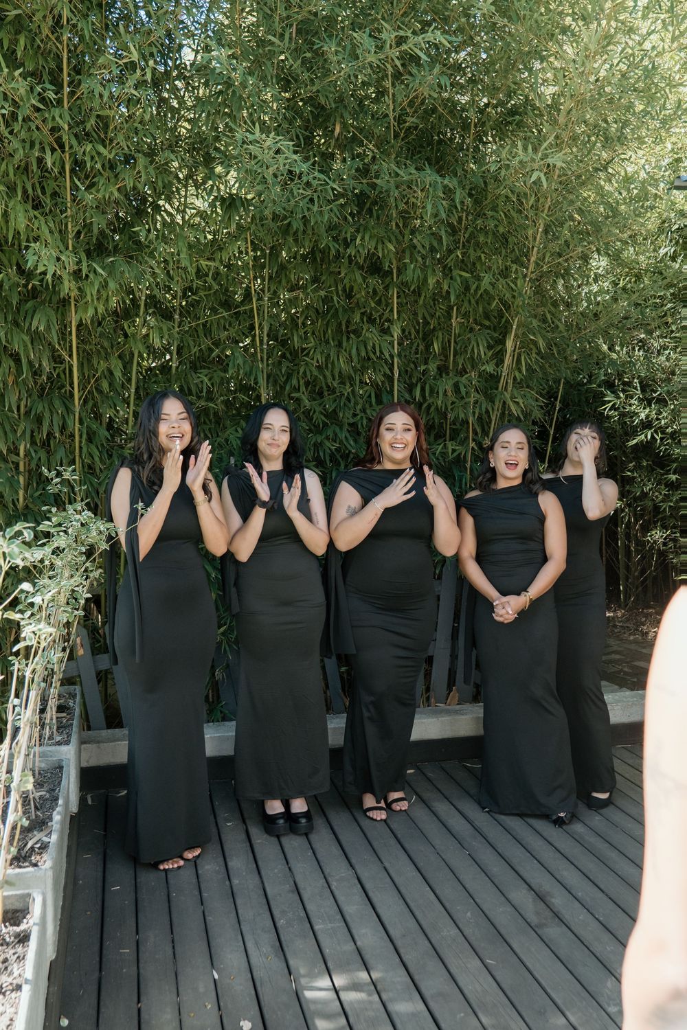 A group in matching black formal dresses pose together on a wooden deck with bamboo plants in the background.