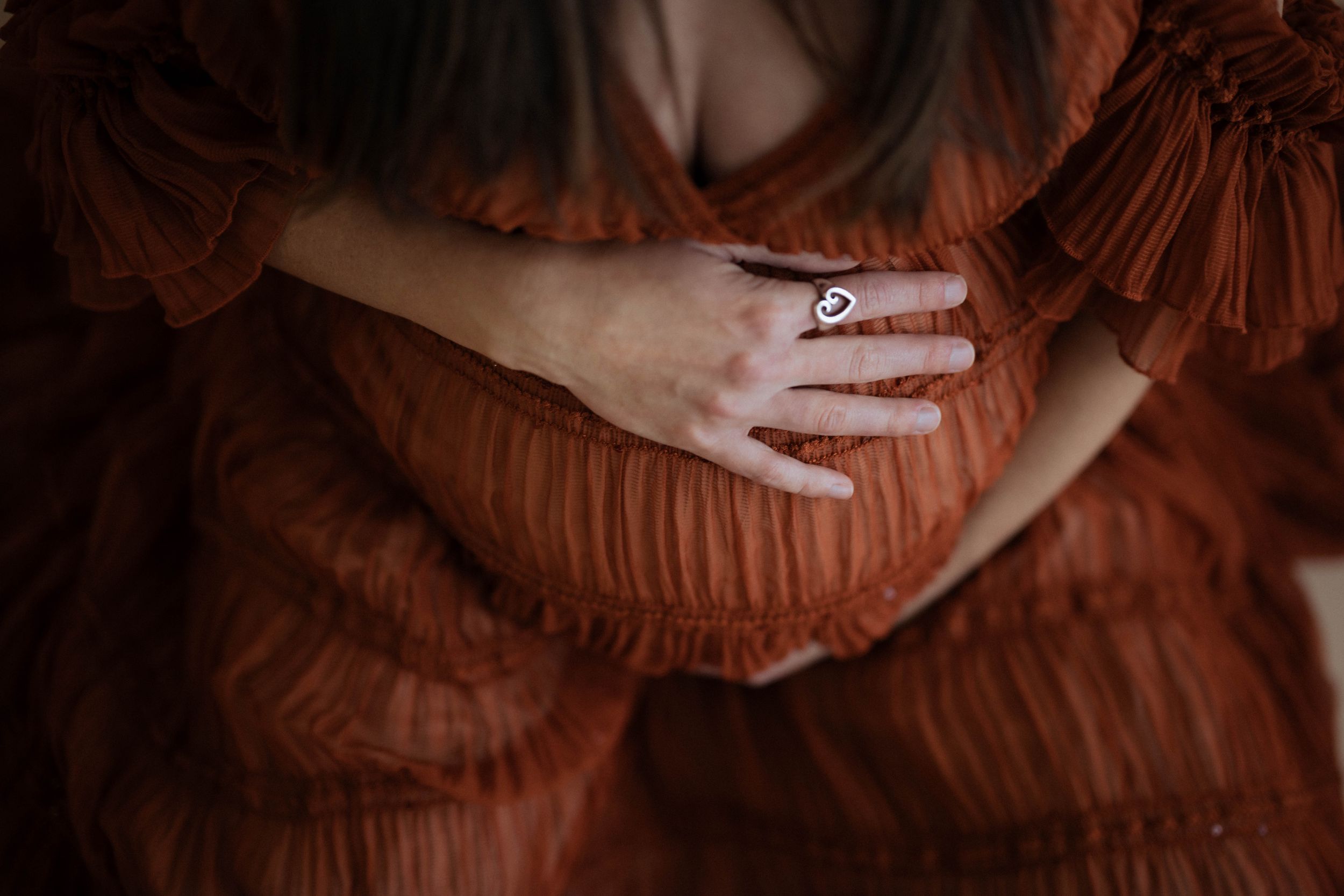 A delicate hand wearing a ring rests on a rust-colored pleated dress in a moody atmospheric portrait.