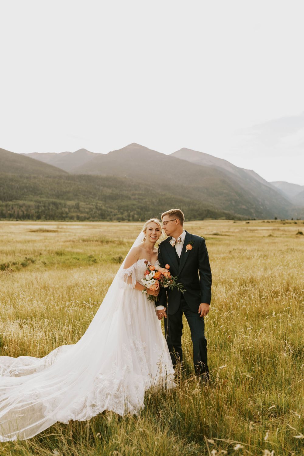 Couple embraces in an open mountain meadow at sunset with a flowing white wedding dress and wildflowers.