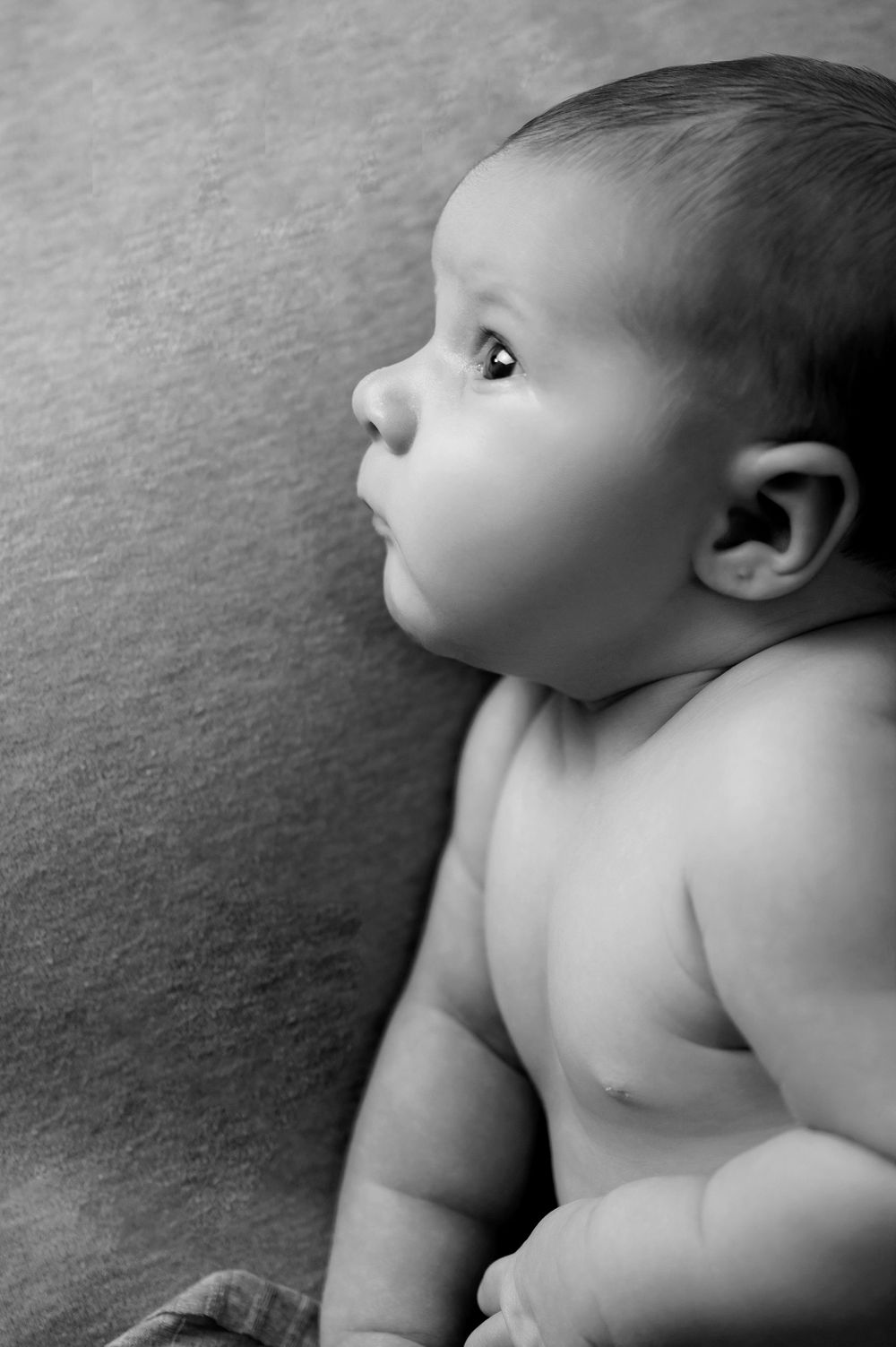A black and white profile shot of an infant with chubby cheeks gazing upward while lying on a textured surface.
