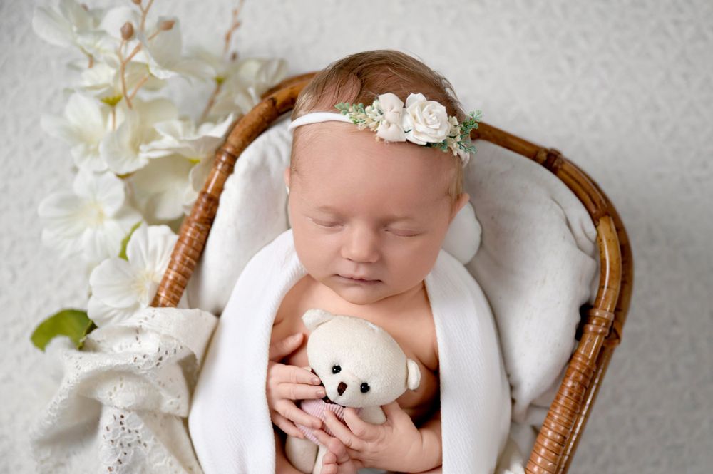 Sleeping newborn rests peacefully in wicker basket with white floral headband and plush panda toy.