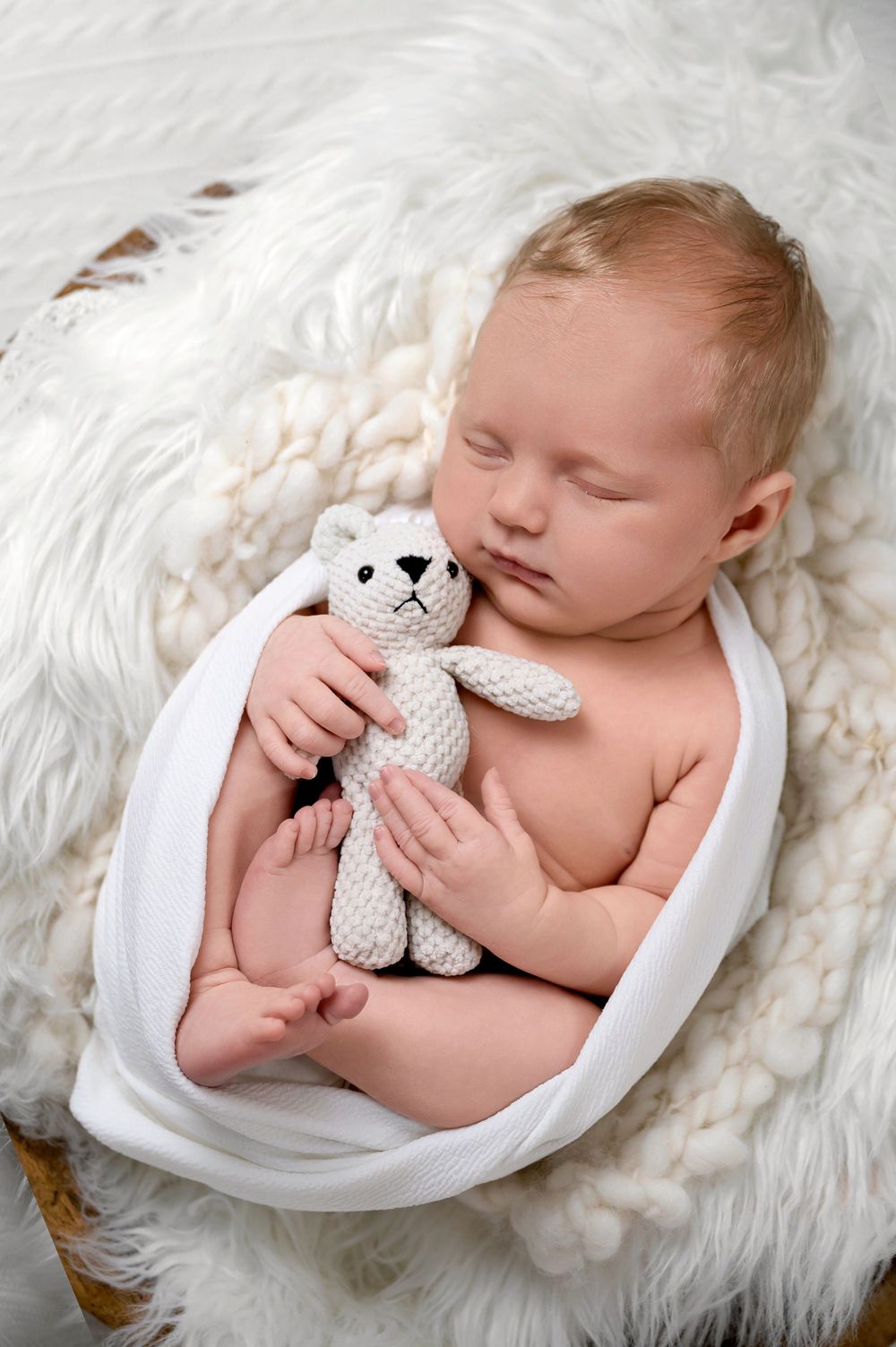 Sleeping newborn cradled in white fabric holding small knitted teddy bear on fluffy white blanket.