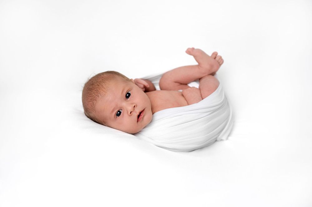 A newborn baby lies swaddled in a white blanket against a white background.
