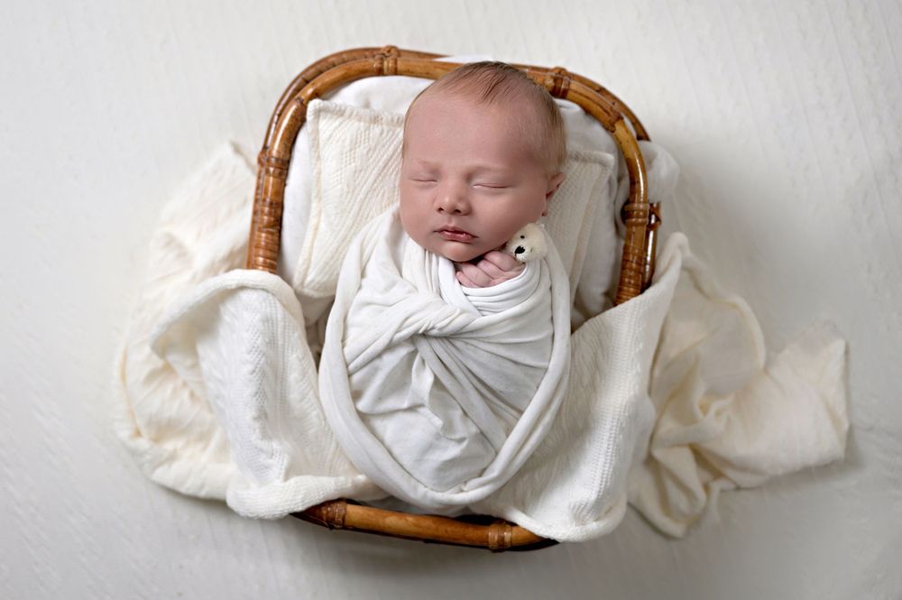 A sleeping newborn wrapped in white fabric rests peacefully in a woven brown basket.