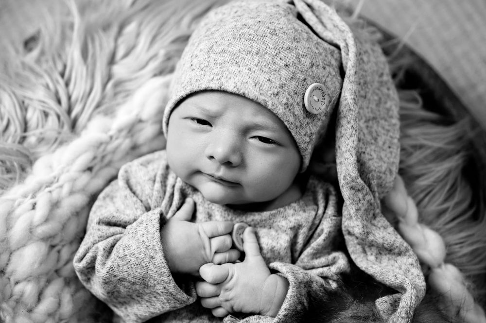 Black and white portrait of infant wearing soft gray knit hat lying on textured blanket.