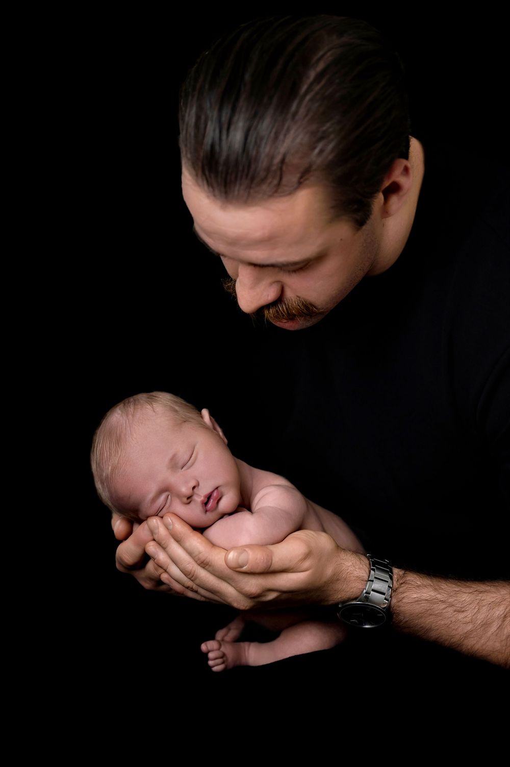 A tender moment captured between father and newborn baby against black background as dad cradles infant in his hands.