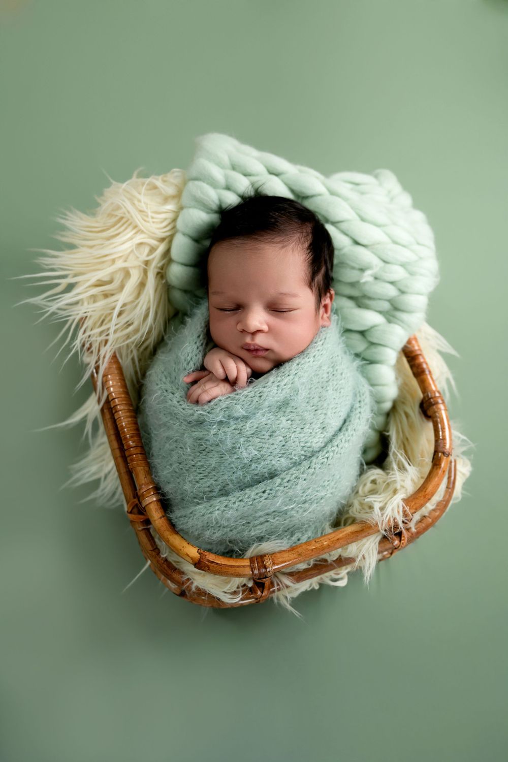 Peaceful sleeping newborn wrapped in mint green swaddle resting in wooden basket with fluffy cream fur lining on green backdrop.