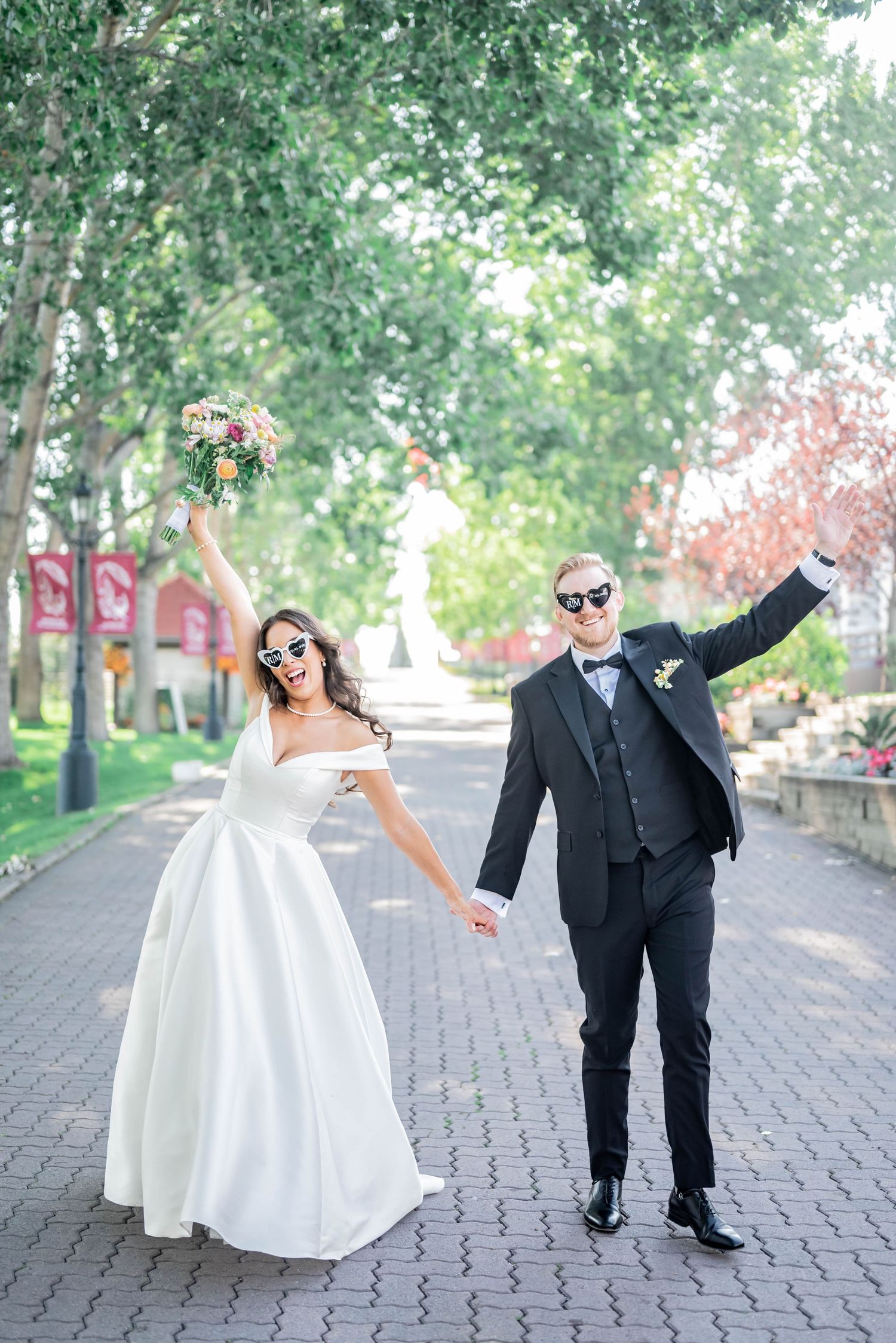 Newlyweds celebrate joyfully with outstretched arms on a tree-lined brick pathway during their wedding photo session.