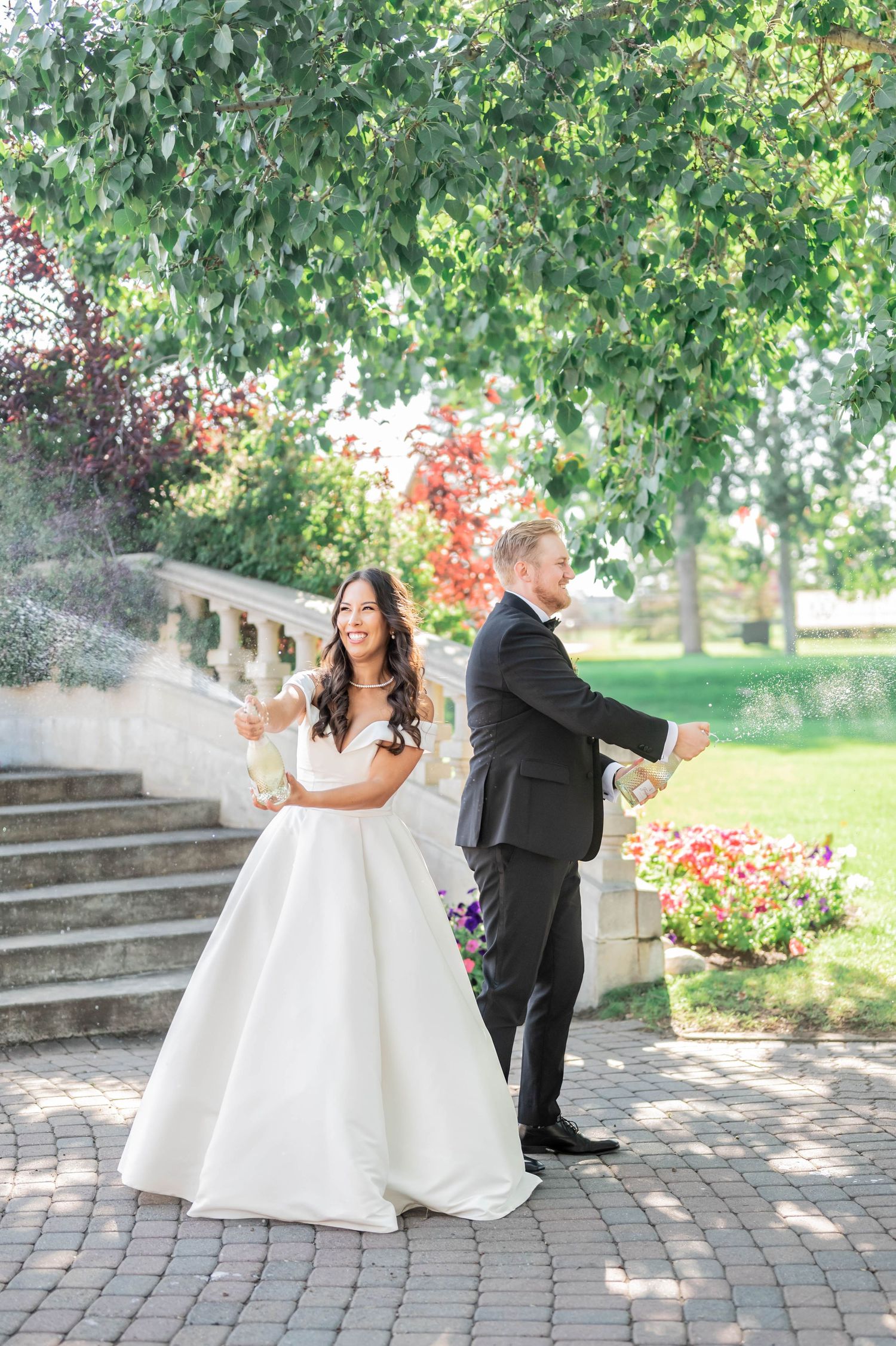 A couple in wedding attire stands back-to-back on a brick patio with stone steps and greenery in the background.