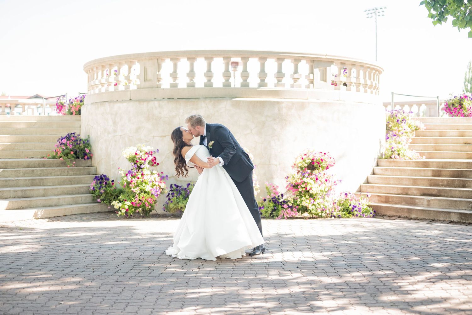 A romantic moment between newlyweds on elegant stone stairs adorned with vibrant floral arrangements in sunlight.