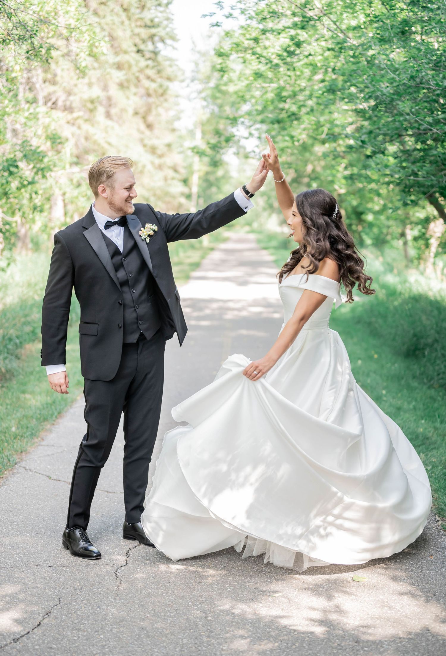 Joyful couple dancing and twirling together on a tree-lined path during their wedding celebration.