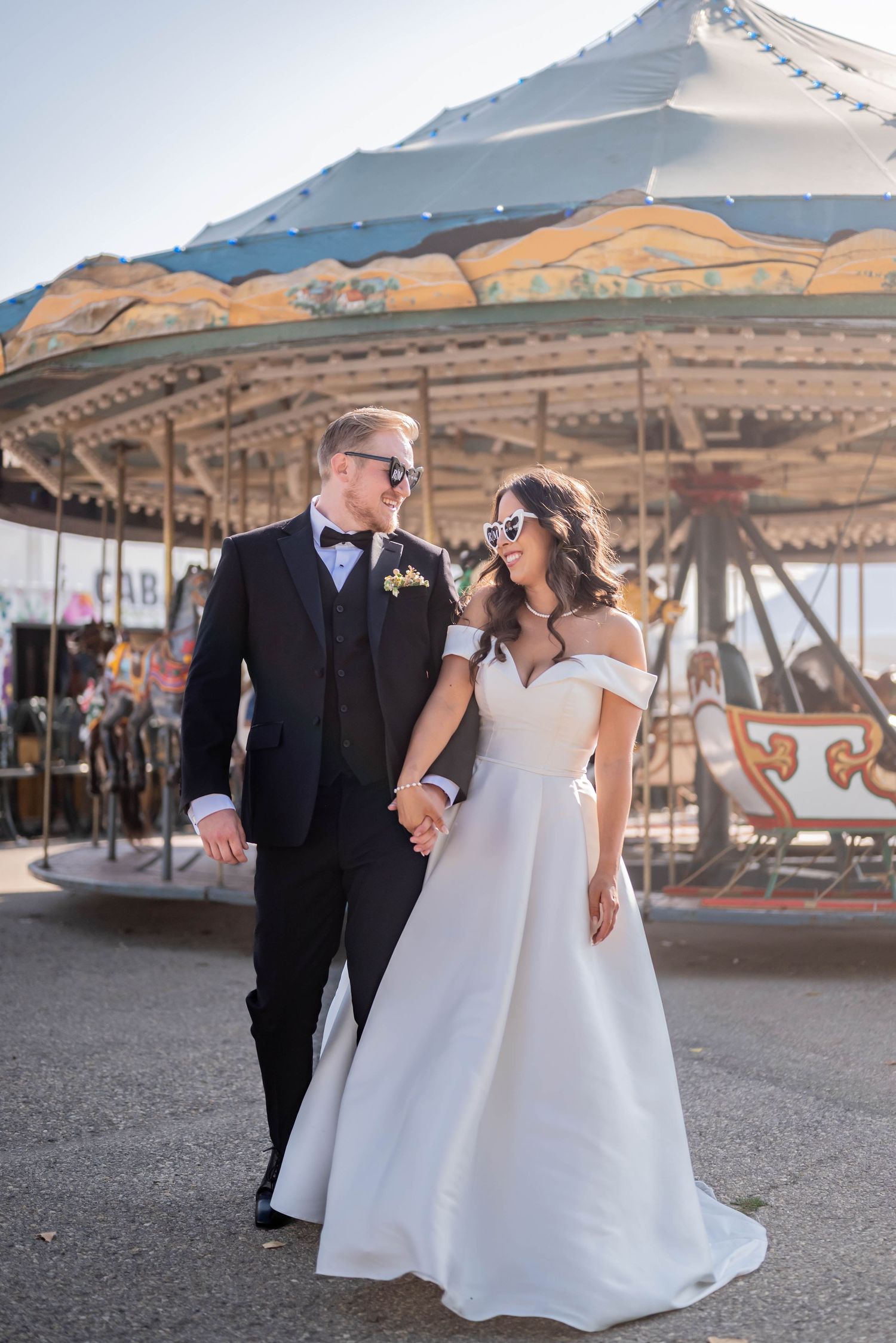 A newlywed couple holds hands and smiles near a vintage carousel at their wedding venue.