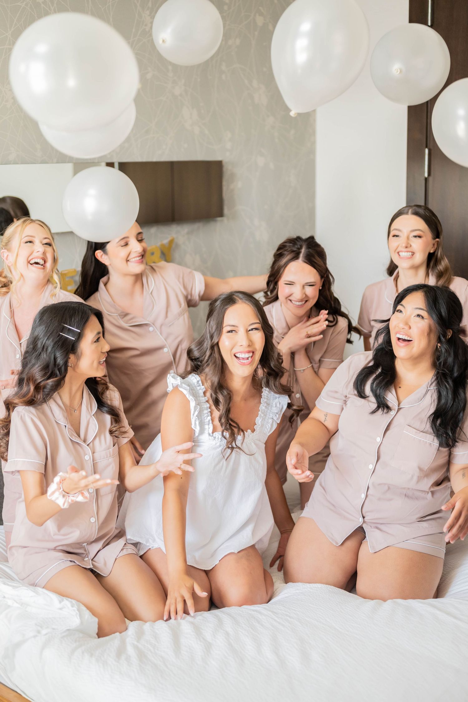 A group in matching light colored sleepwear lounges on a bed with white balloons during a celebration party.