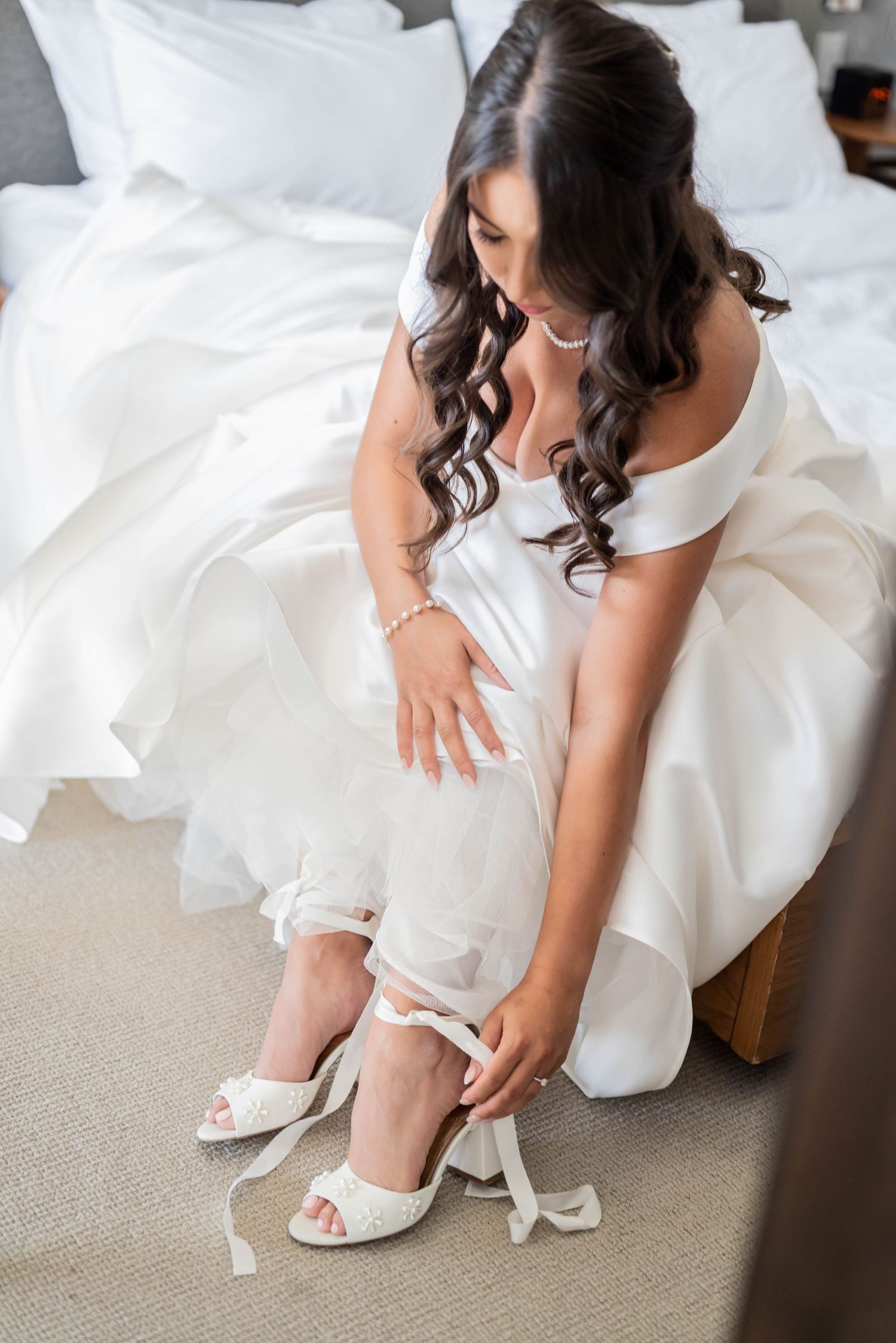 A bride in a white wedding dress sits on a bed while putting on elegant white heeled sandals.
