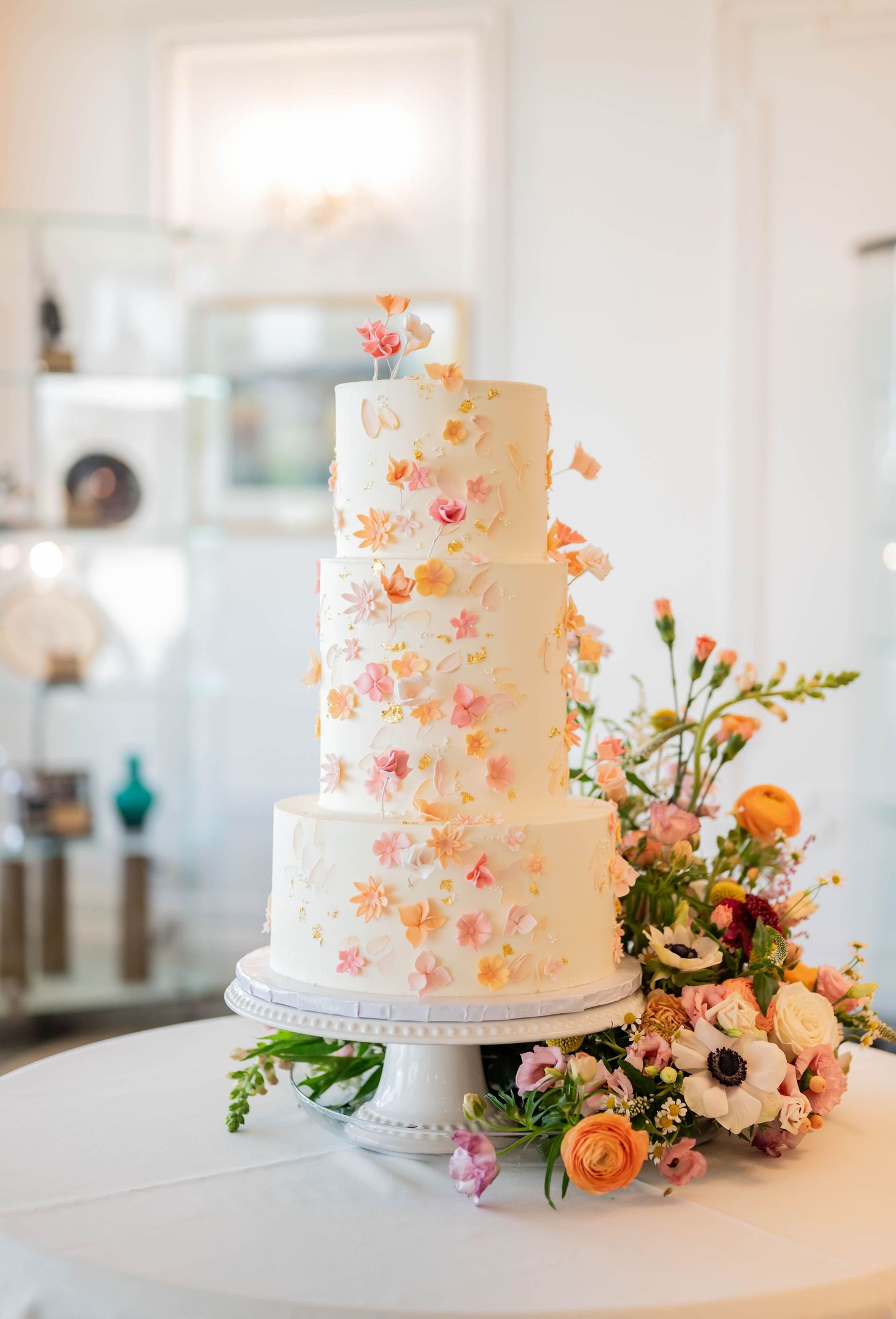 Elegant four-tier white wedding cake decorated with delicate coral flowers and greenery on a white cake stand.
