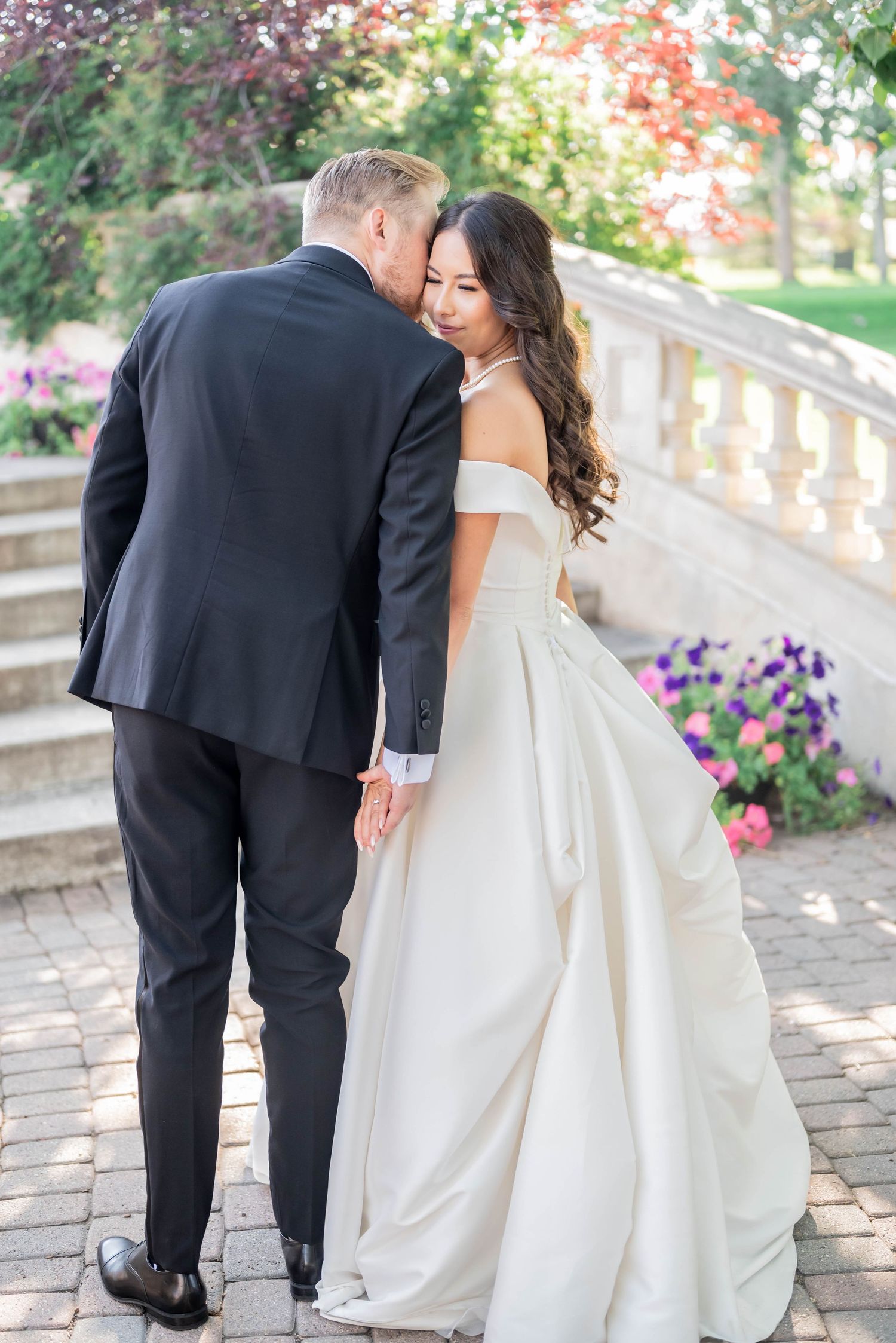 Loving couple in wedding attire embraces on elegant stone steps surrounded by vibrant purple and pink flowers.