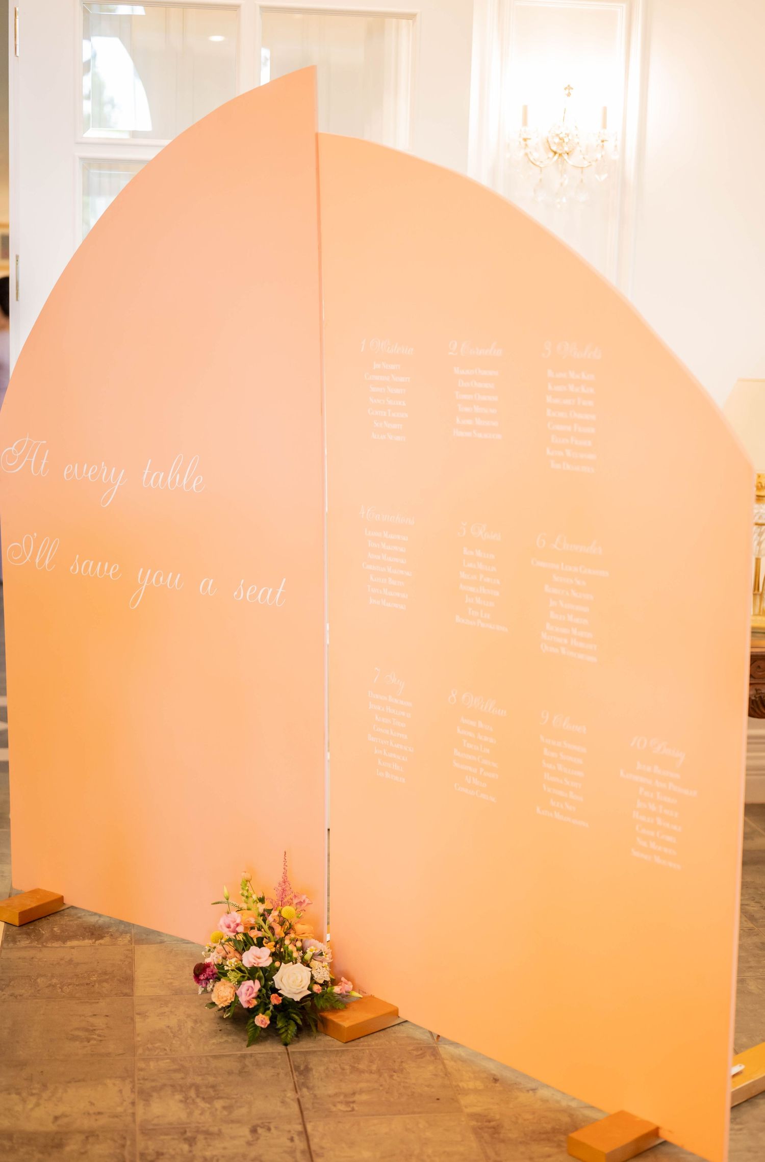 A peach-colored curved wedding seating chart display with a floral arrangement at its base.