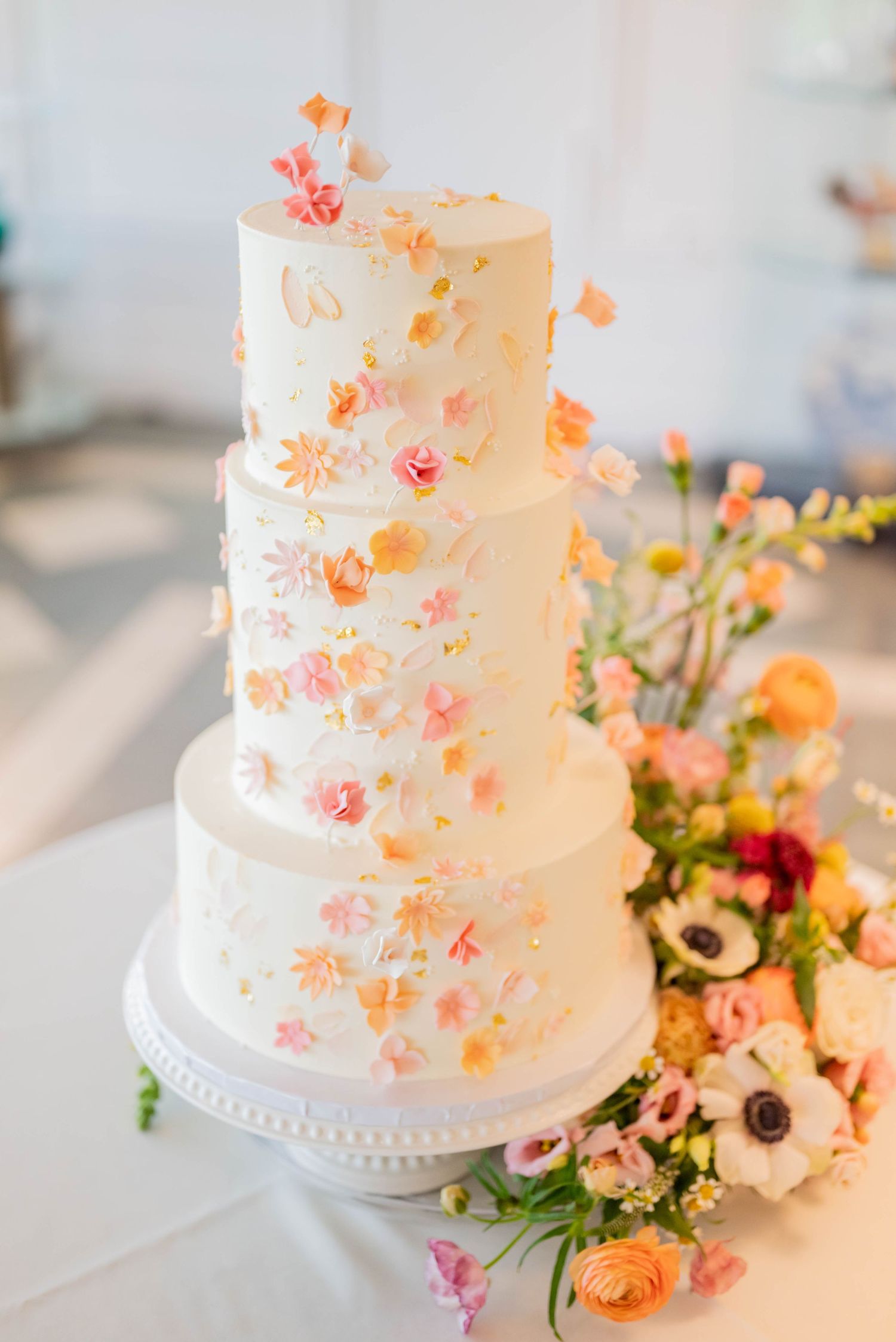 Four-tier white wedding cake decorated with delicate peach and coral sugar flowers on a crystal stand with floral accents.