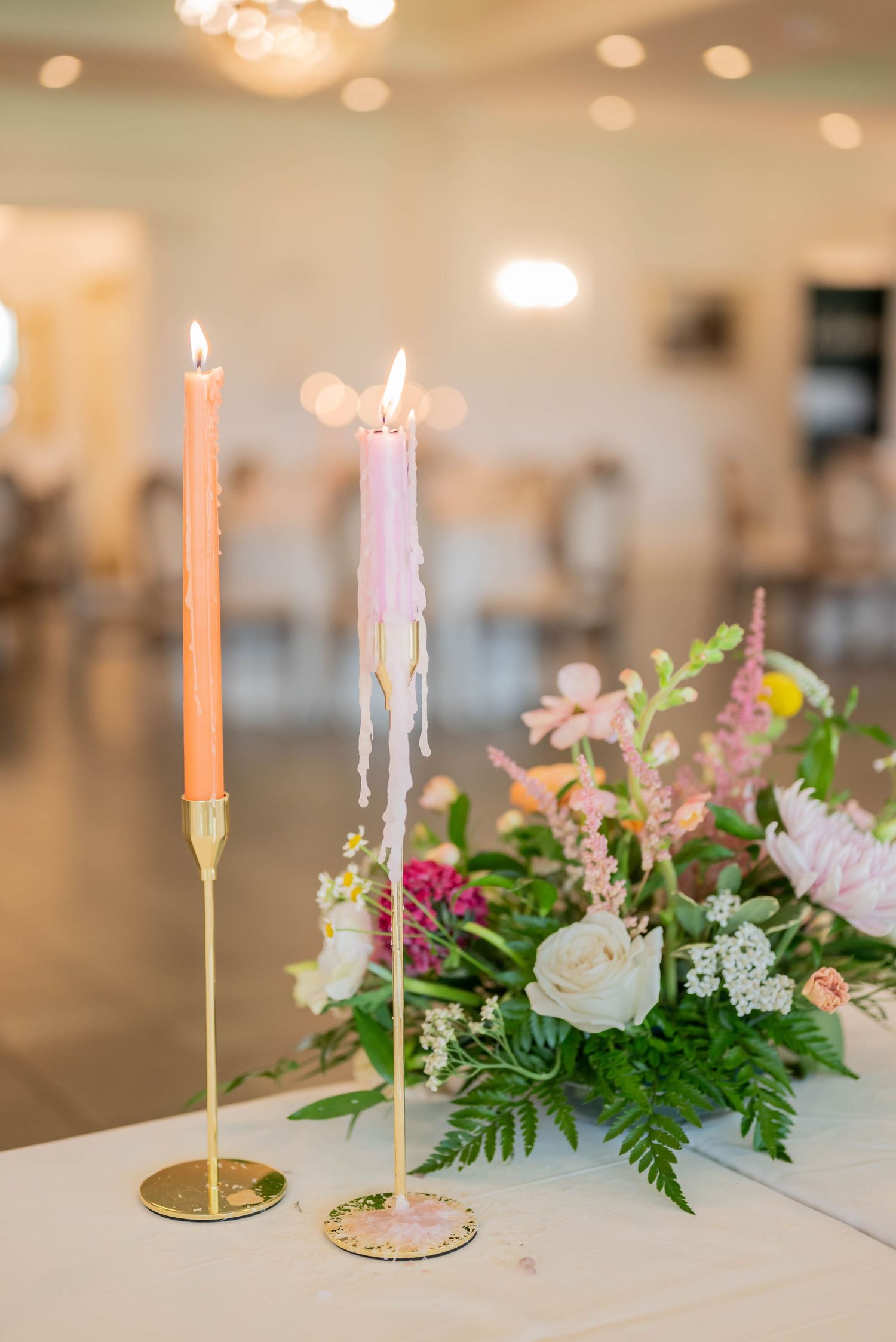 Lit taper candles in gold holders beside pink and white floral centerpiece arrangement at an elegant event venue.