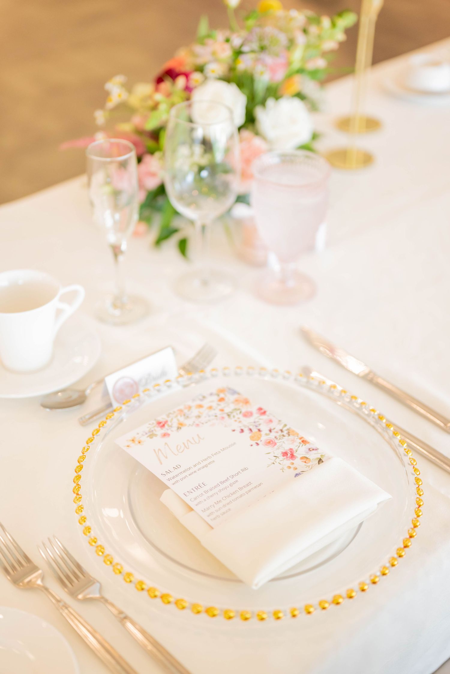 Elegant place setting with gold-rimmed charger plate, floral menu card, and soft pink floral centerpiece on white tablecloth.