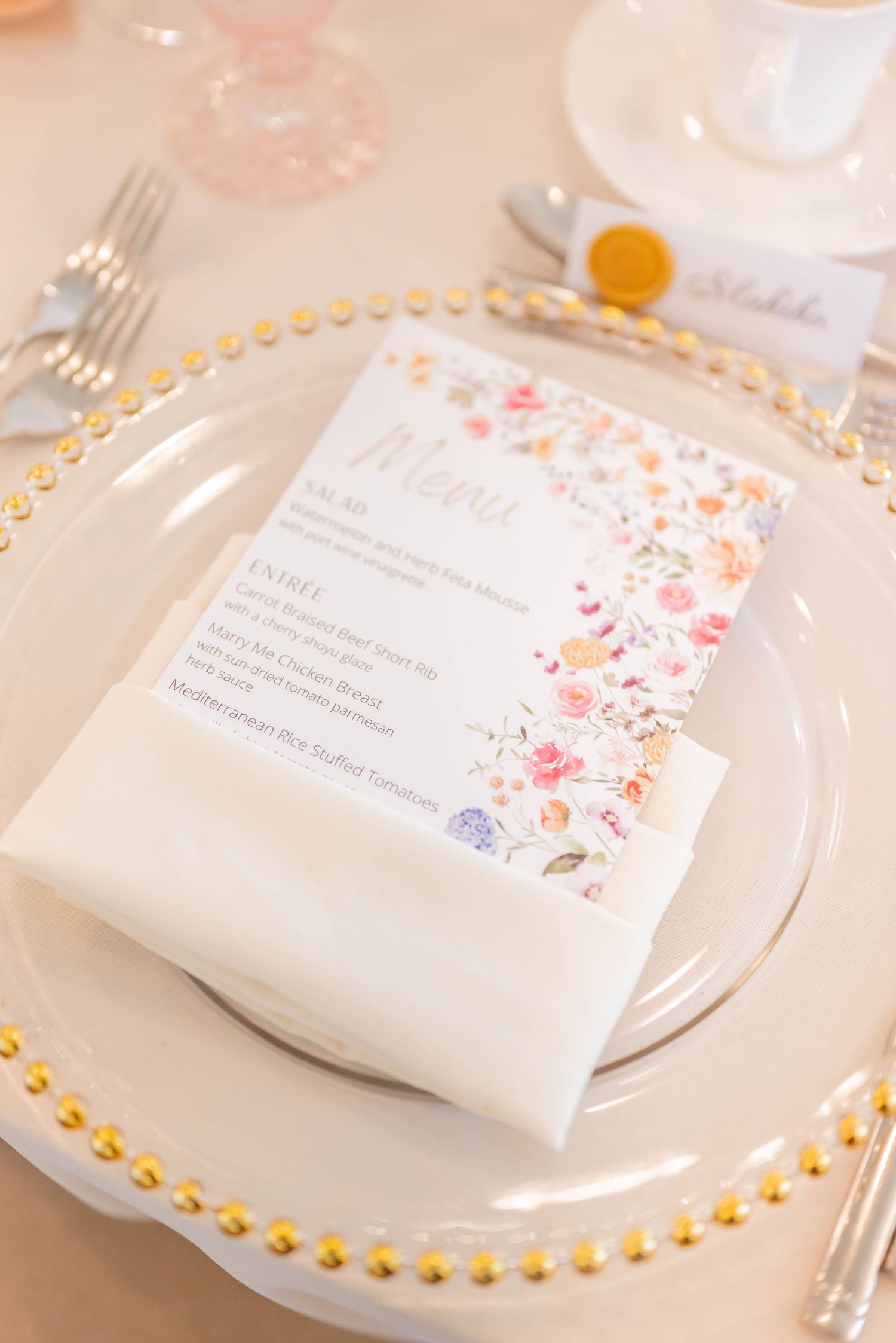 Wedding place settings featuring gold-rimmed glass charger plates and floral decorated menu cards on white tablecloths.