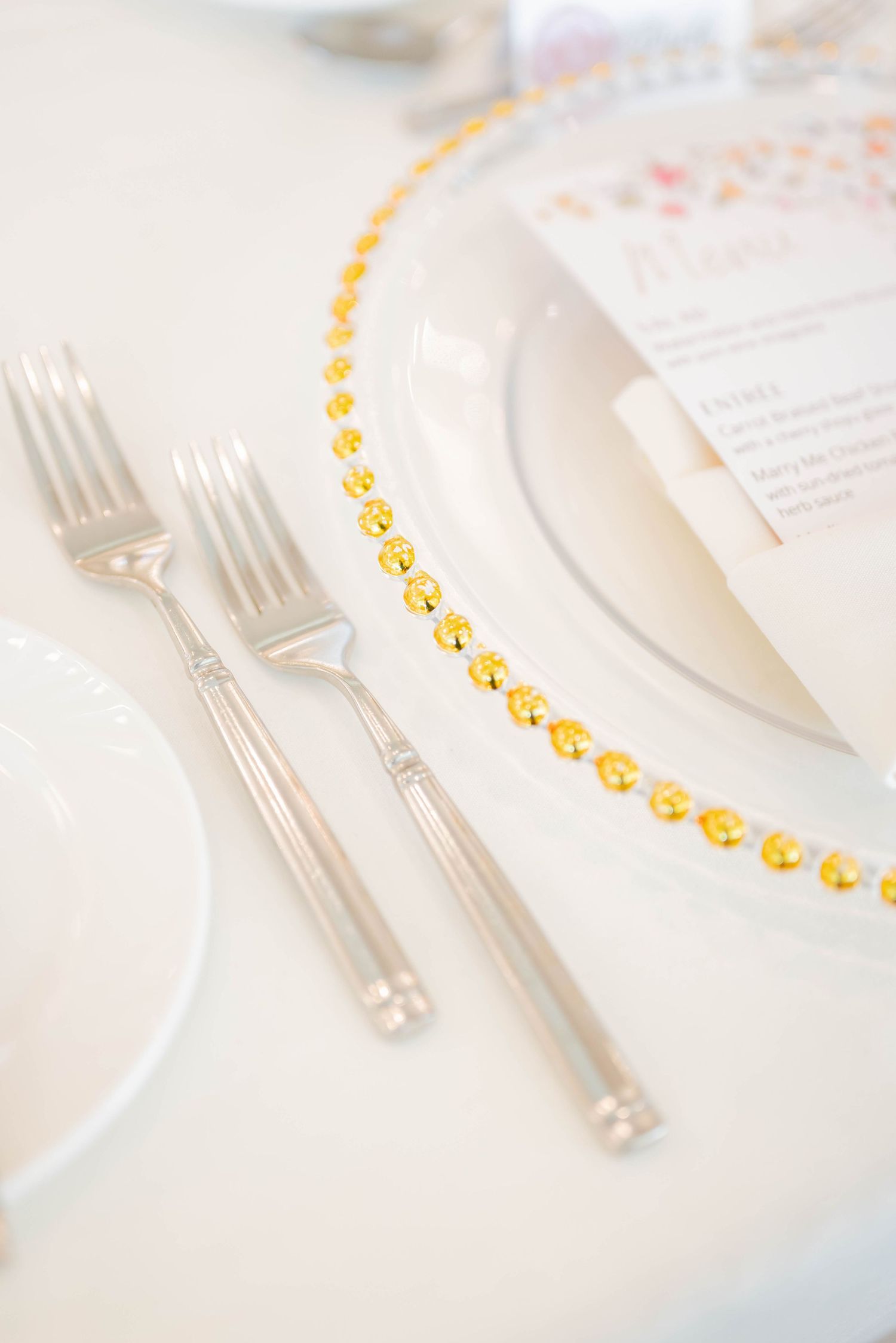 Elegant place setting with silver forks and gold beaded trim on white tablecloth for formal dining.