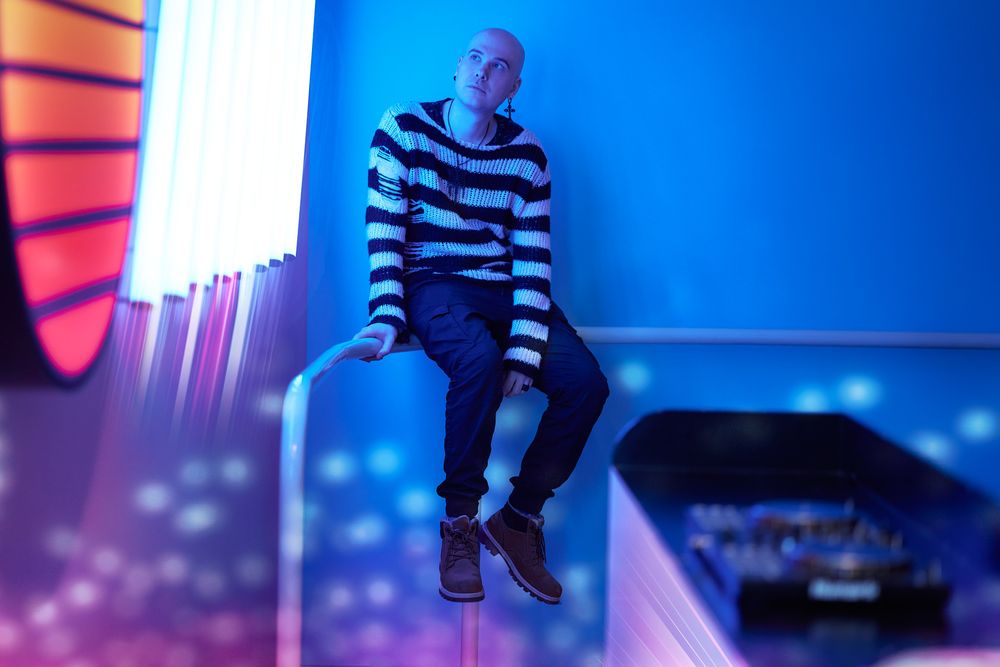 Person in striped clothing sits in a blue-lit room with multiple screens and modern gaming equipment visible.