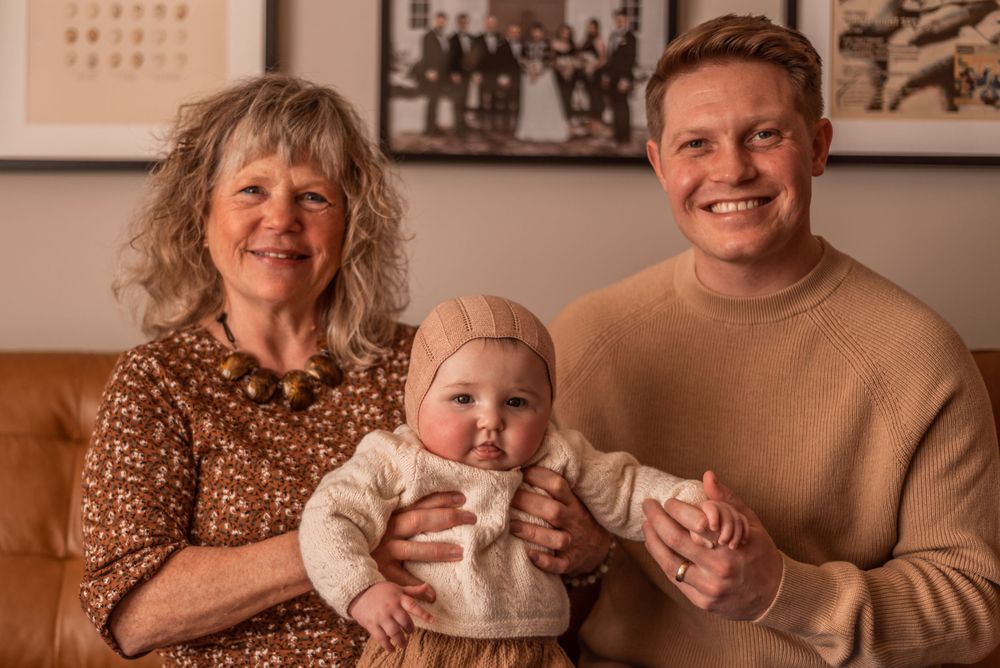 Three generations pose together in a cozy living room setting wearing warm-toned clothing.