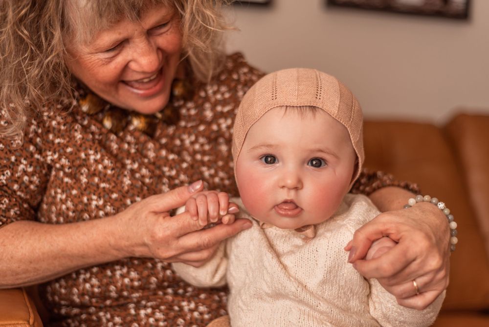 Family members smile while holding a baby wearing a cream knit outfit and beige cap.