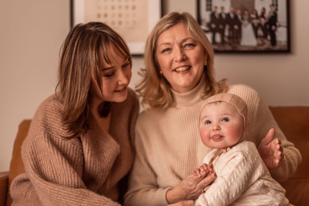 Three generations share loving smiles while holding a baby in cream-colored clothing.