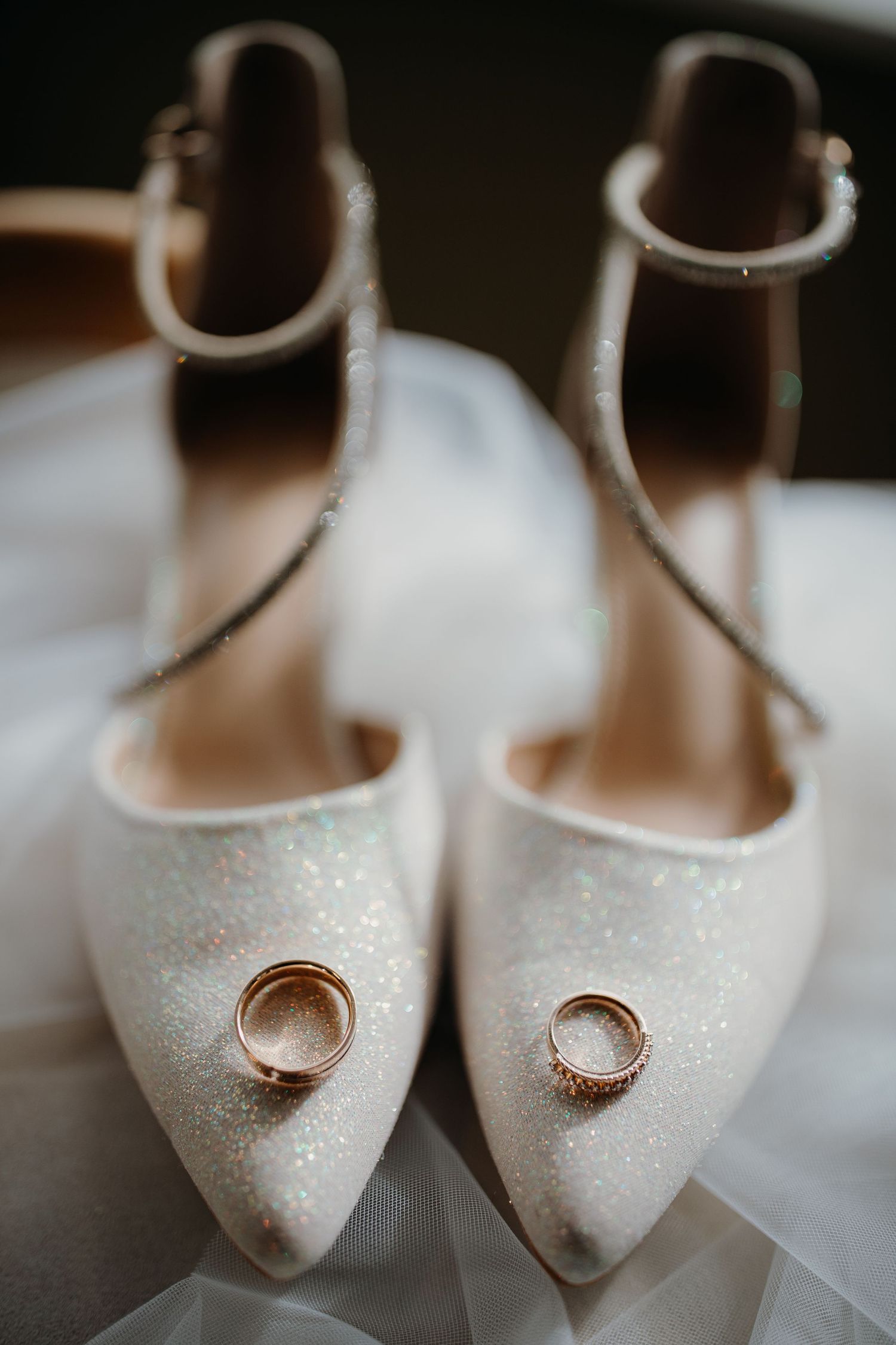 Wedding rings displayed artistically on white glittery bridal shoes with ankle straps and pointed toes.