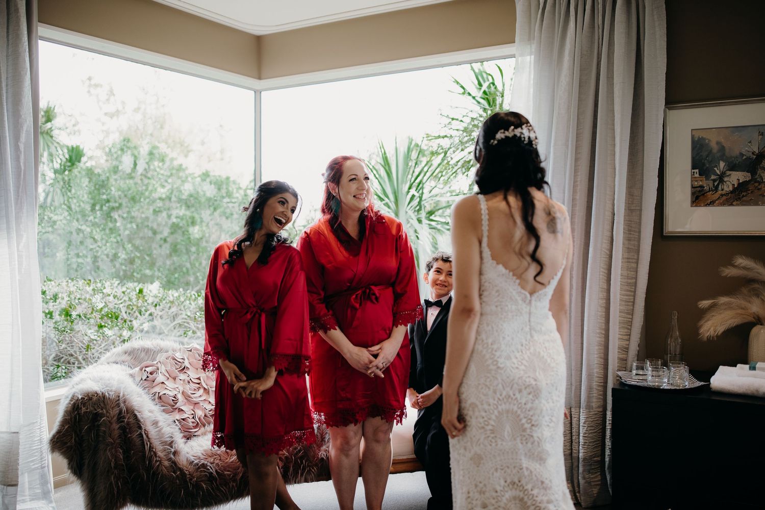 Bridesmaids in matching red robes stand with young ring bearer before bride in elegant white wedding gown.
