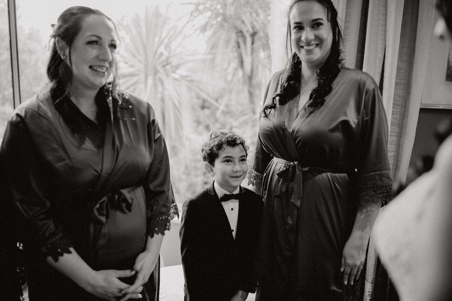 A black and white photo shows two women in dark dresses standing with a young child in a tuxedo and bow tie.