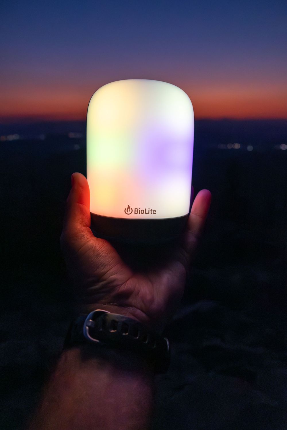 Glowing multicolor BioLite lantern held up against purple sunset sky.