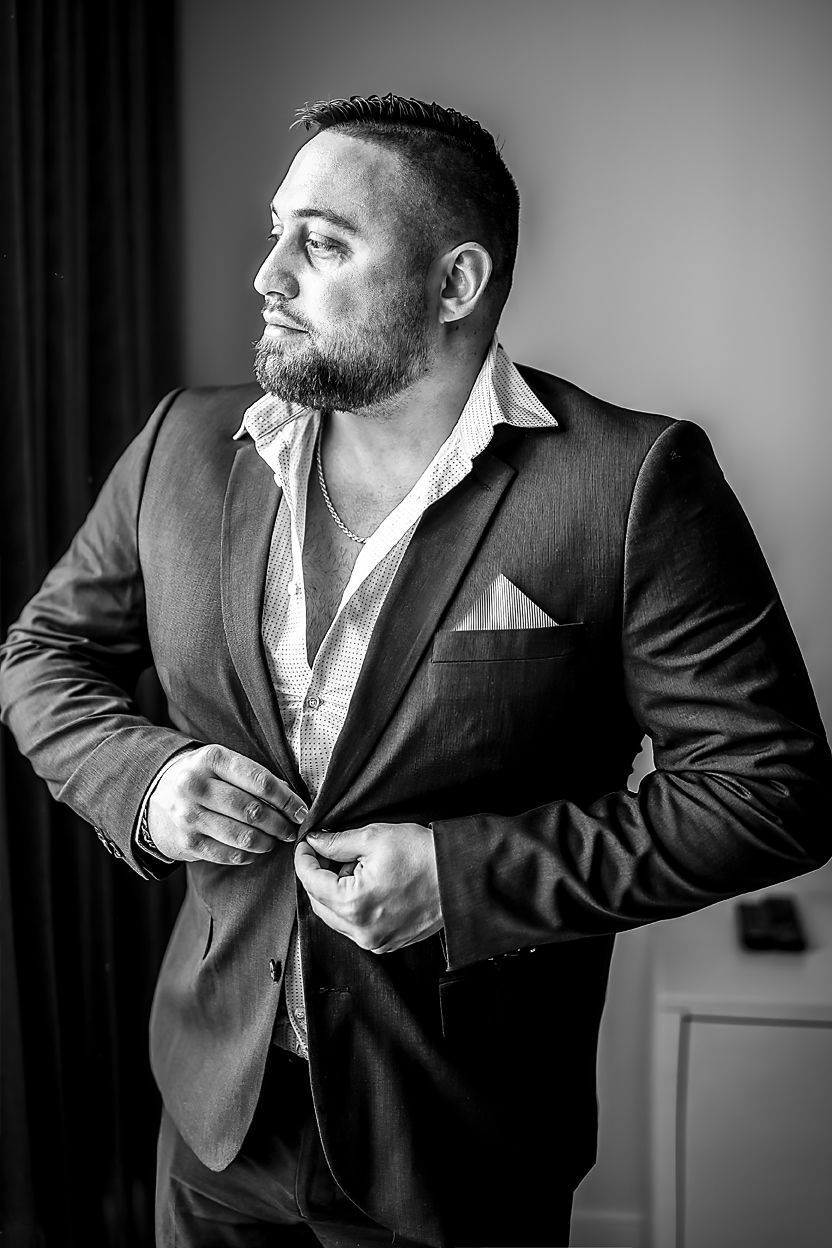 Black and white portrait of a bearded man in a dark suit jacket and open white dress shirt looking to the side.