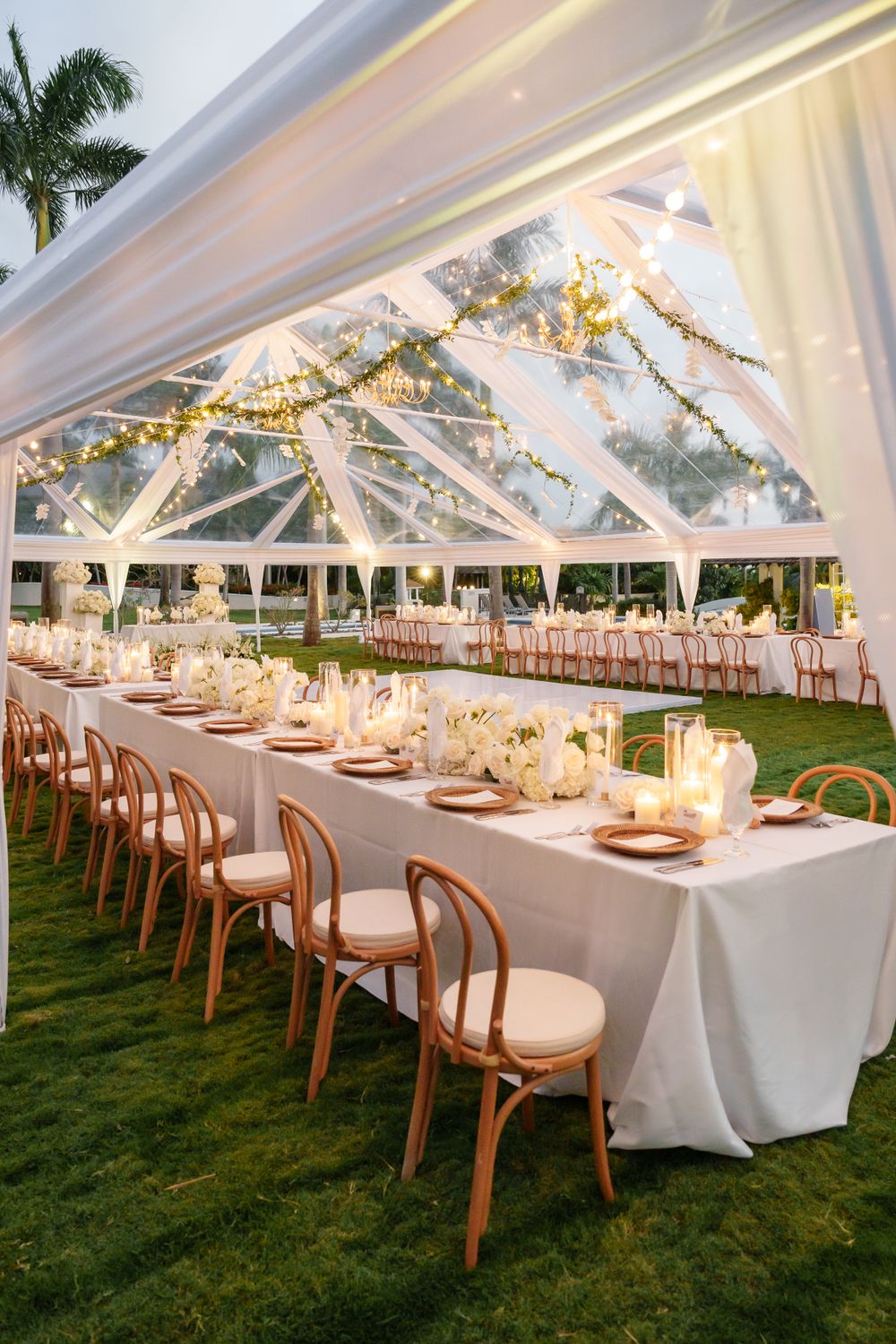Elegant outdoor reception with long white tables under a clear tent decorated with string lights and floral arrangements.