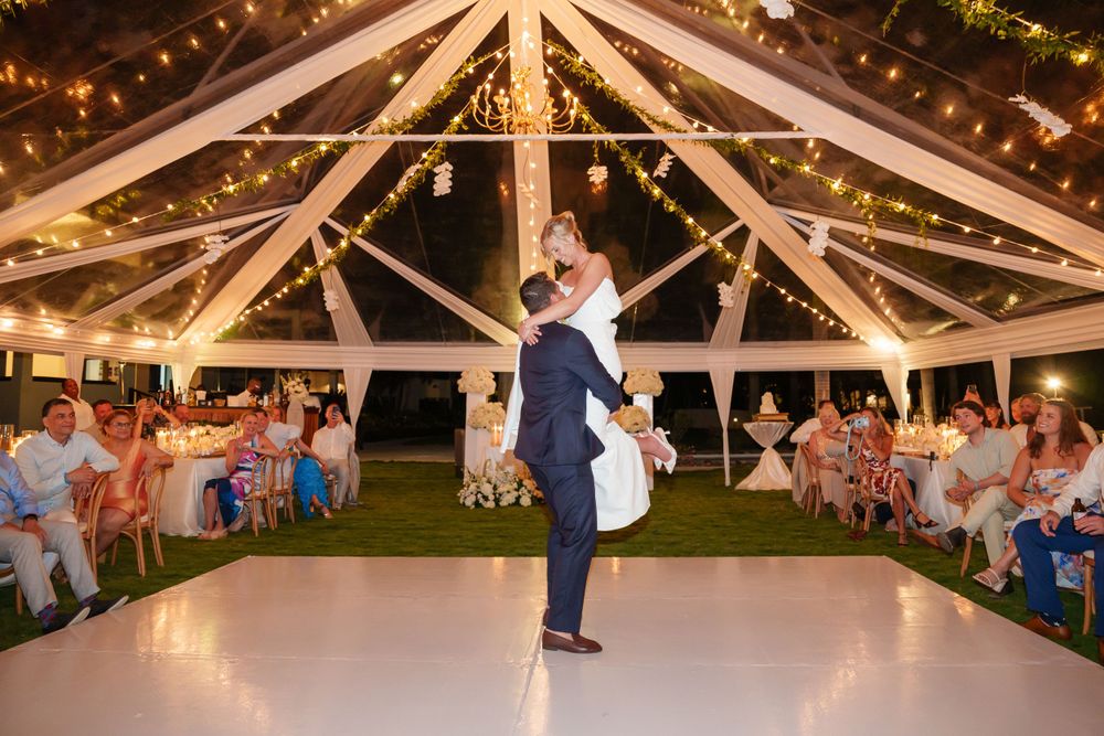A newlywed couple dances under a white tent structure with string lights and watching guests.